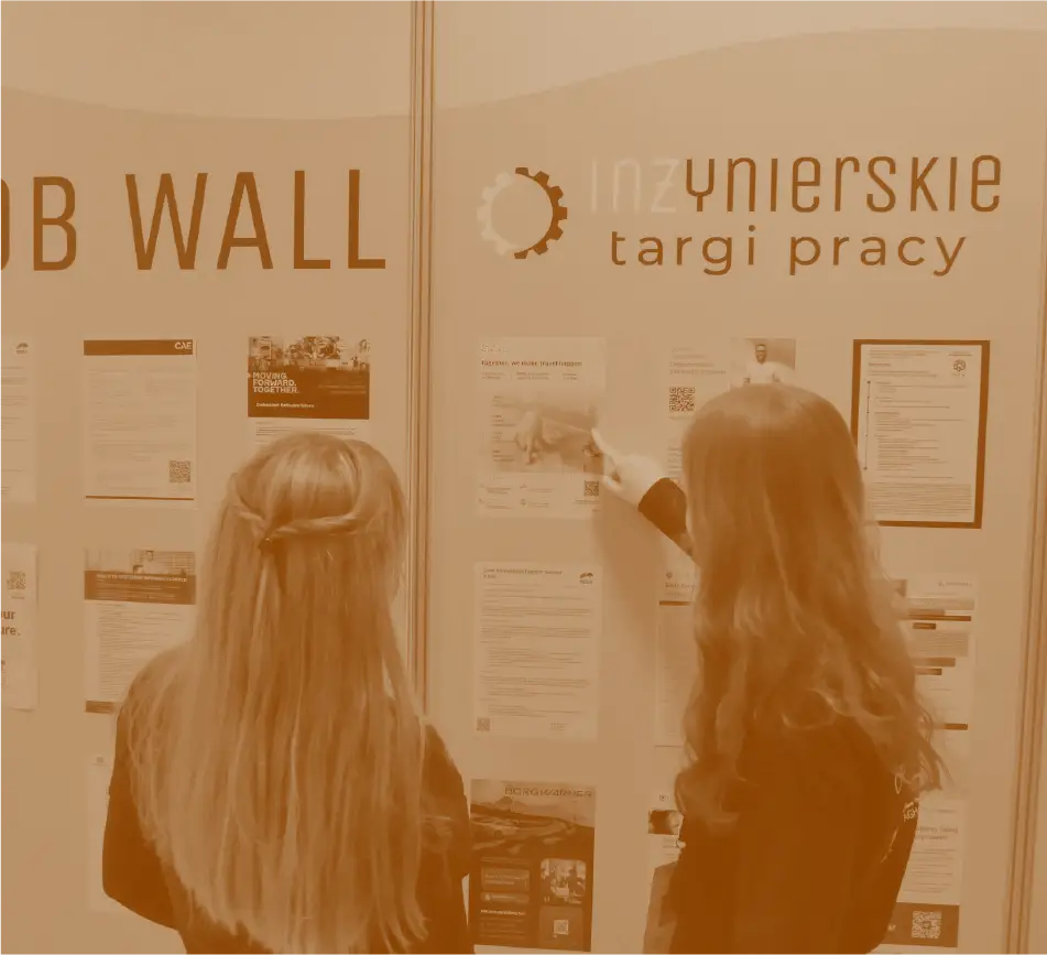 Dwie kobiety stoją tyłem i przeglądają ogłoszenia o pracę zawieszone na ściance z napisem „JOB WALL” oraz logo „Inżynierskie Targi Pracy”.
