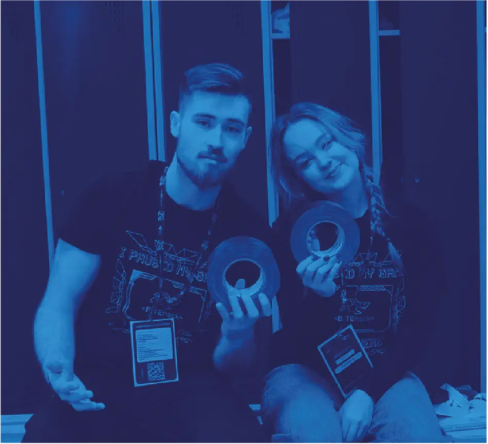 Portret dwójki uśmiechniętych organizatorów BITEhack (BEST AGH Kraków) trzymających rolki taśmy montażowej.